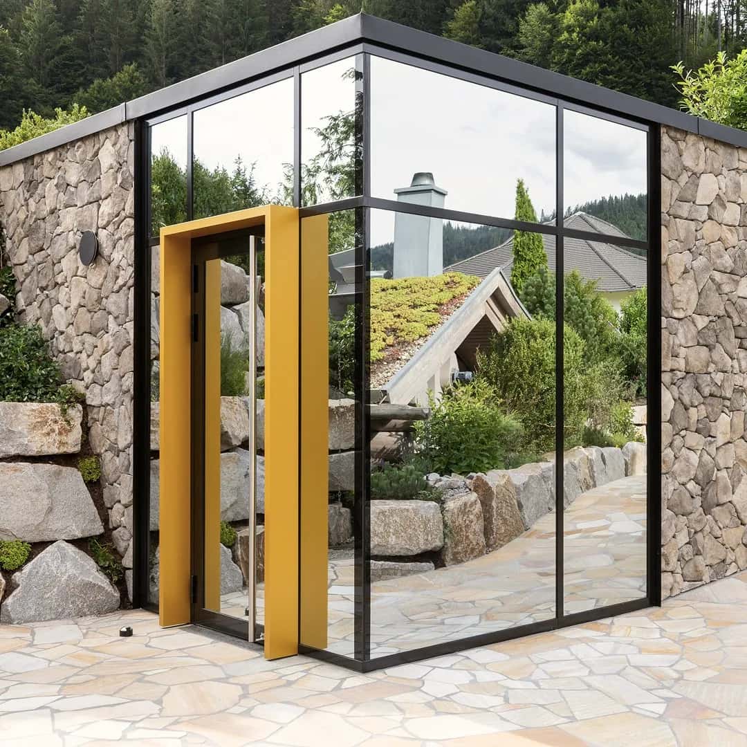 Moderne Sauna mit großflächiger Glasfront, schwarzem Rahmen und gelber Eingangstür, eingebettet in eine Natursteinmauer und umgeben von üppigem Grün.