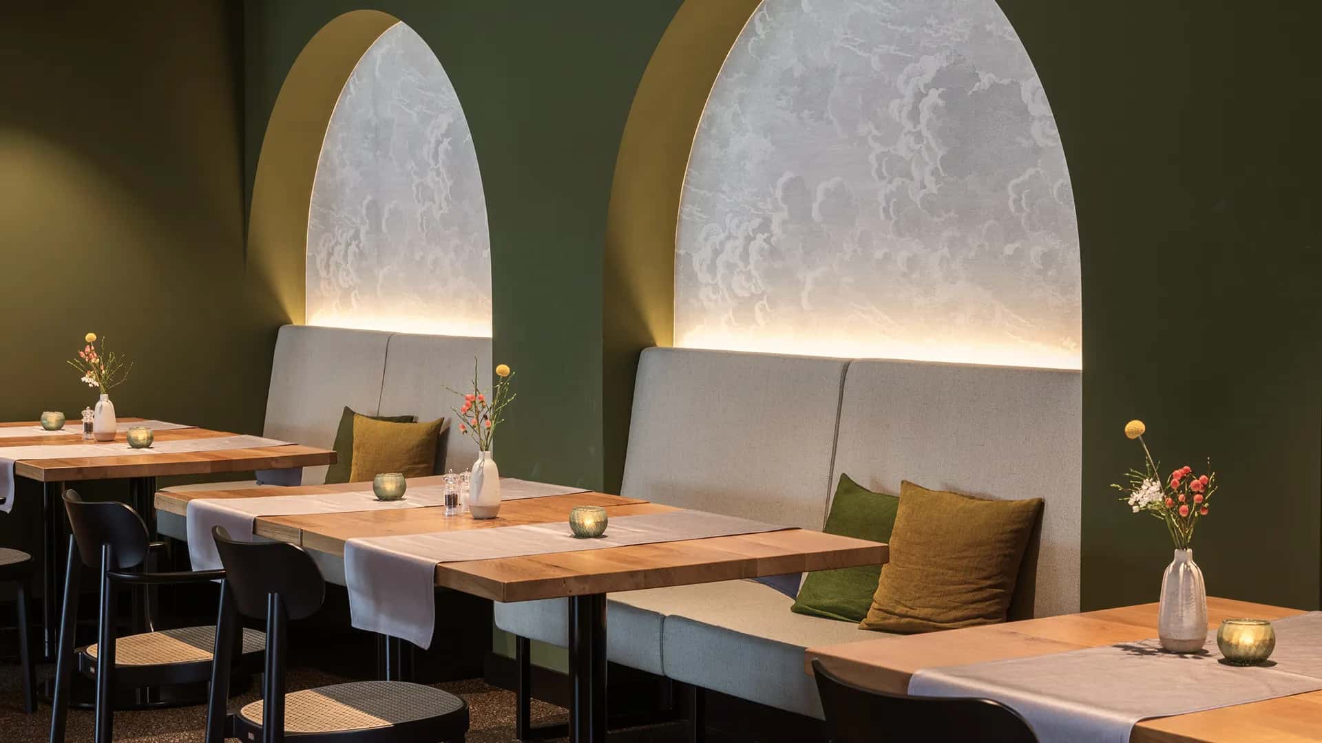 Restaurantbereich mit olivgrüner Wand, hinterleuchteten Bogennischen und hellen Holztischen.