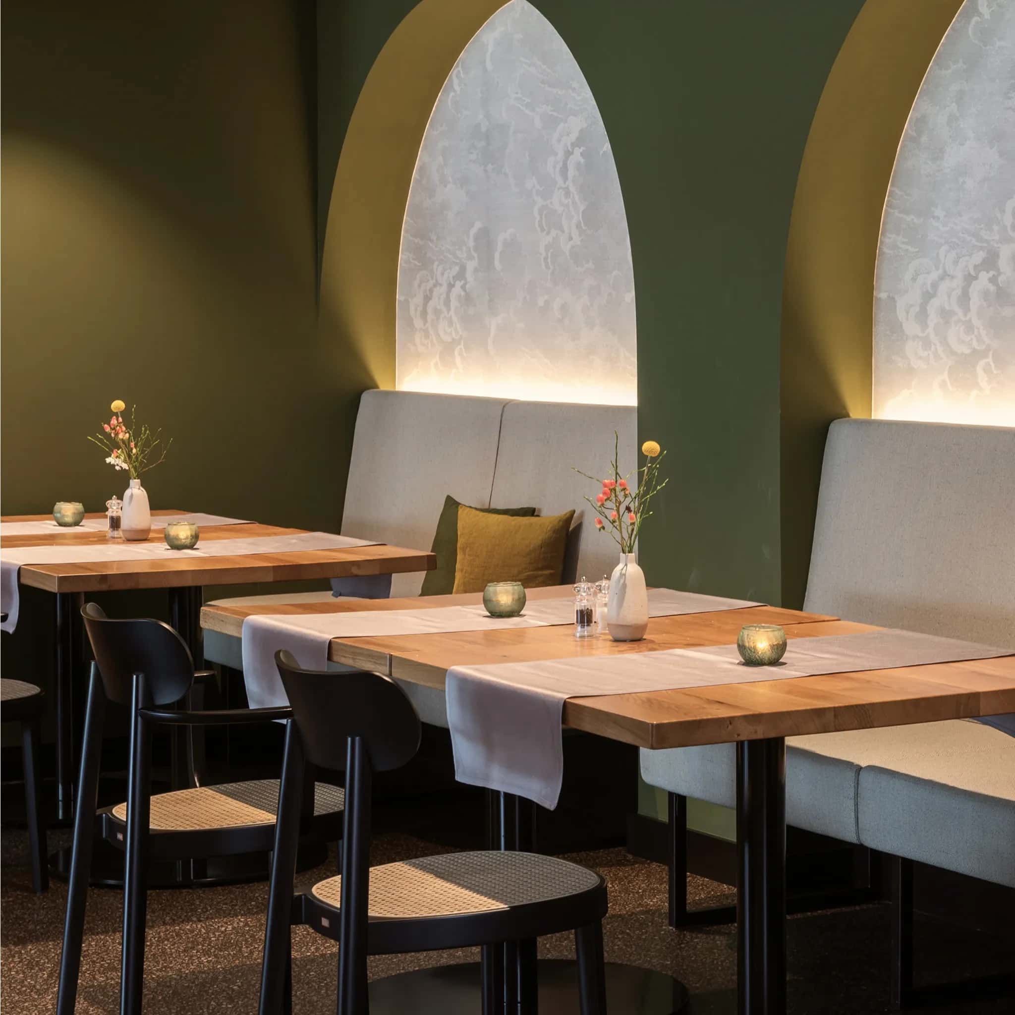 Restaurantbereich mit olivgrüner Wand, hinterleuchteten Bogennischen und hellen Holztischen.